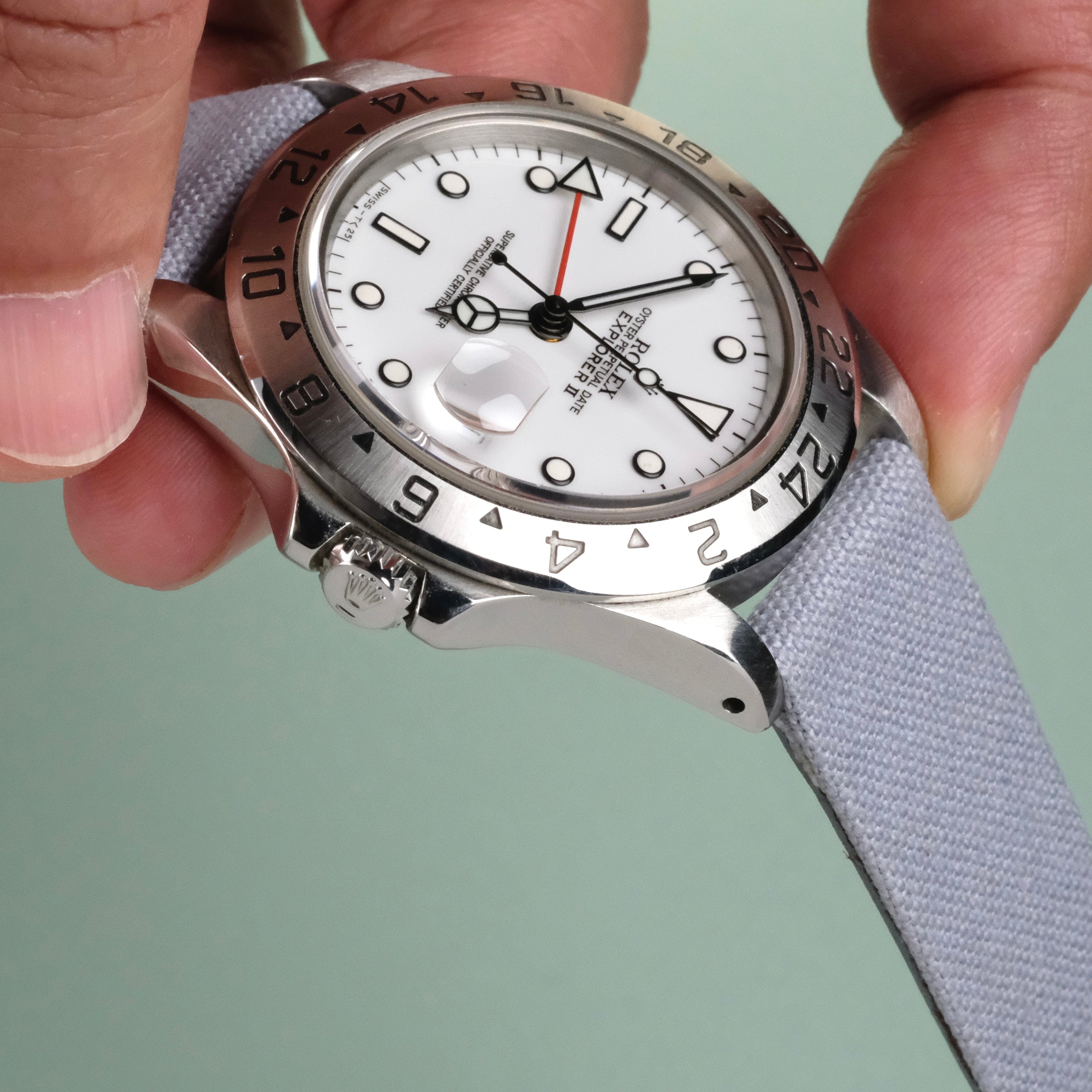 A hand holds a silver wristwatch with a white dial and black hour markers, fitted with the RSM Watch Strap Fine Canvas Watch Strap in Metallic Blue, against a pale green background. The round bezel is engraved with numbers.