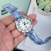 A hand holds a wristwatch with a white dial, silver bezel, and an RSM Watch Strap Ocean Camo Adjustable Single Pass Watch Strap with stainless steel hardware; a succulent plant and white tray are in the background.