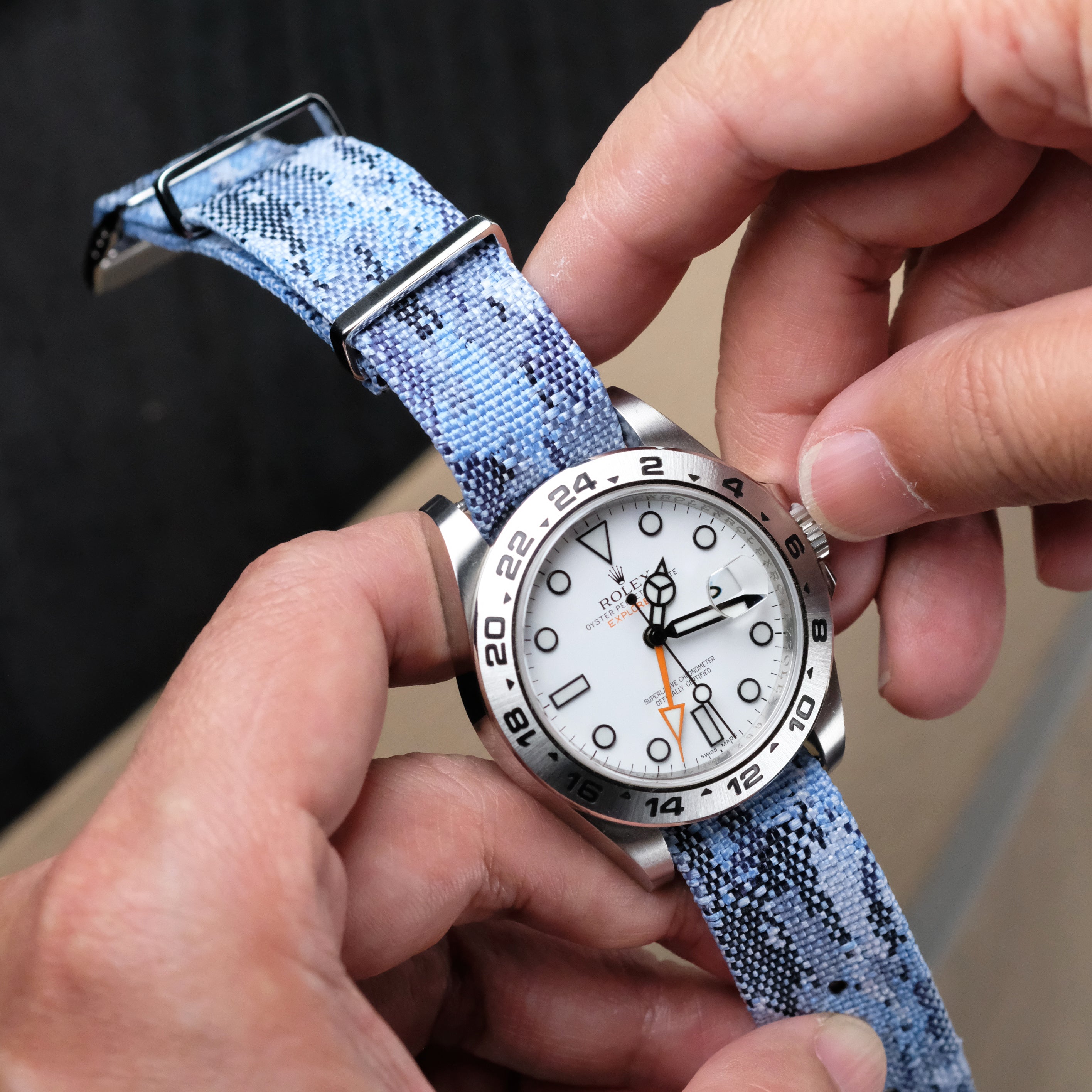 A person adjusts a wristwatch with a white dial and black markings, fitted with an RSM Watch Strap Ocean Camo Adjustable Single Pass jacquard-woven polyester strap and stainless steel hardware, holding it between both hands.