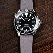 A close-up of an Omega Seamaster with a black wavy dial and silver hands, featuring an RSM Watch Strap Fine Canvas Watch Strap in Mid Grey, placed on a dark wooden surface.