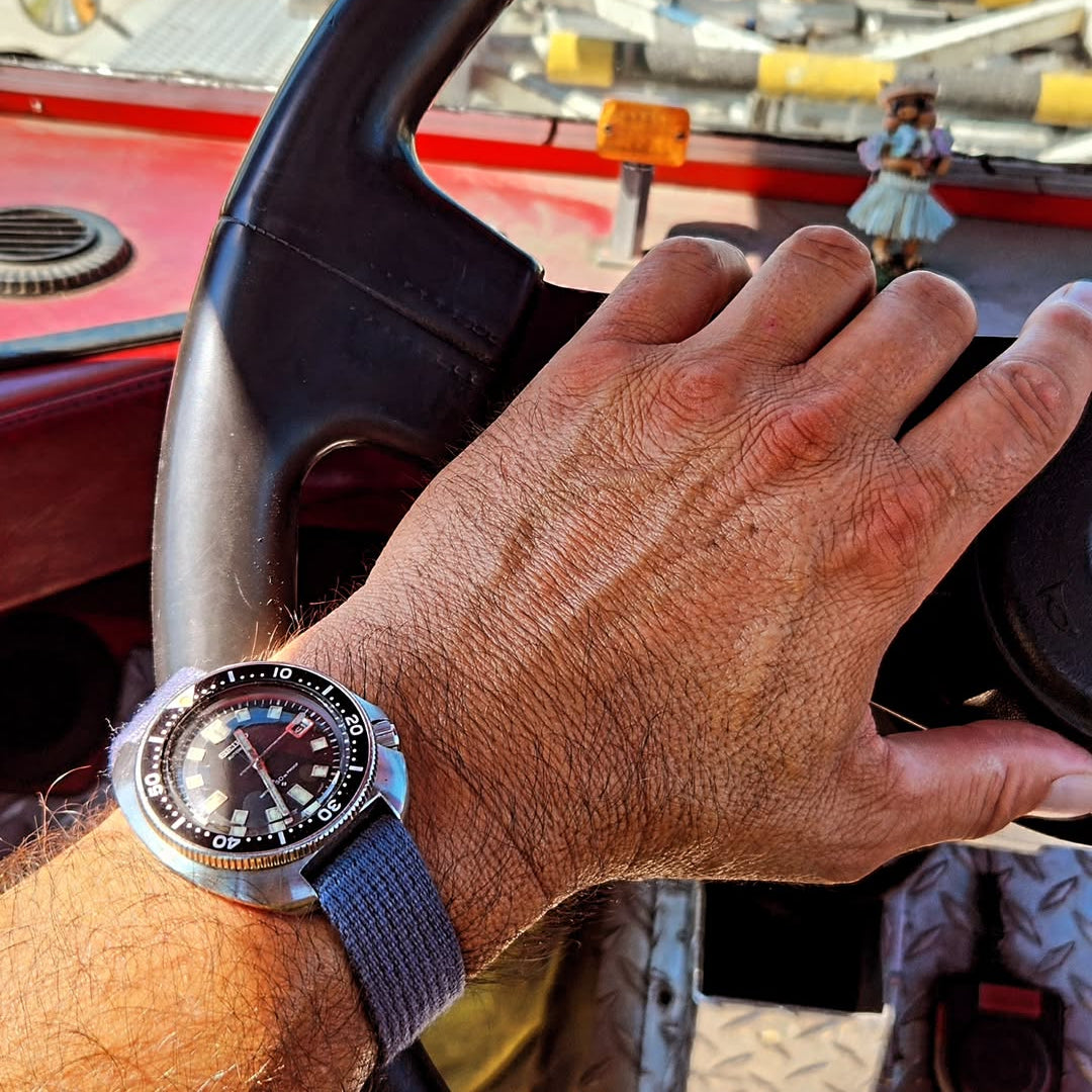 A person wearing the VTG Mil in Blue by RSM with a black dial and rotating bezel holds the steering wheel, showcasing the stylish watch strap.