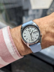 A wrist wearing a Seiko watch with a white grid dial, day-date display, silver case, and an RSM Watch Strap Fine Canvas Watch Strap in Light Blue. The person also wears a pink and white checkered shirt.