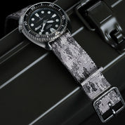 The RSM Watch Strap Snow Camo 2-Piece, a monochrome camo fabric strap, is shown on a black dive watch with a round face, white hour markers, and a rotating bezel, resting atop a dark metal case.