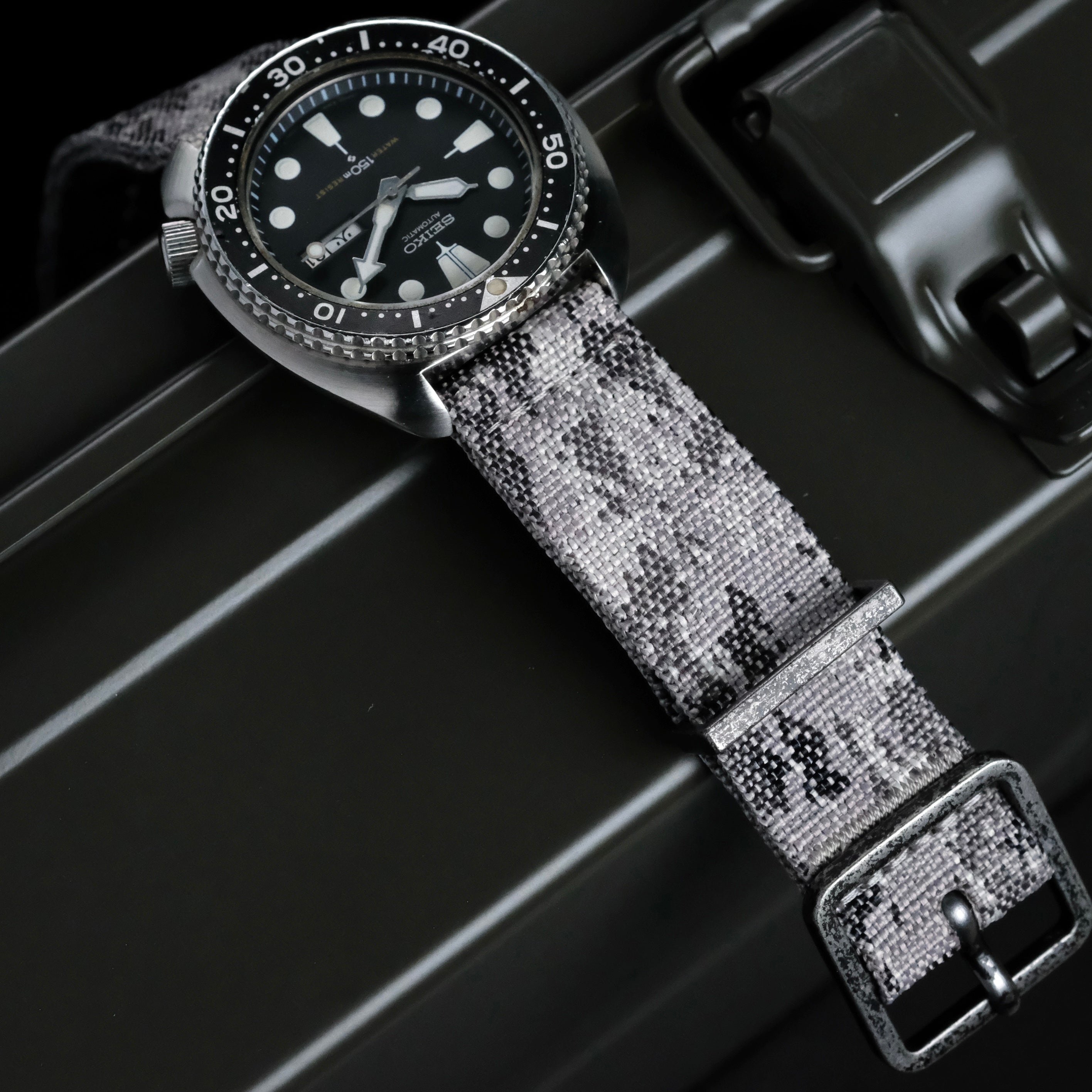 The RSM Watch Strap Snow Camo 2-Piece, a monochrome camo fabric strap, is shown on a black dive watch with a round face, white hour markers, and a rotating bezel, resting atop a dark metal case.