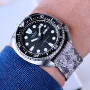 A Seiko dive watch with a black bezel, black dial, and white hour markers is worn on the wrist in a blue sleeve, paired with an RSM Watch Strap Snow Camo 2-Piece jacquard woven strap.