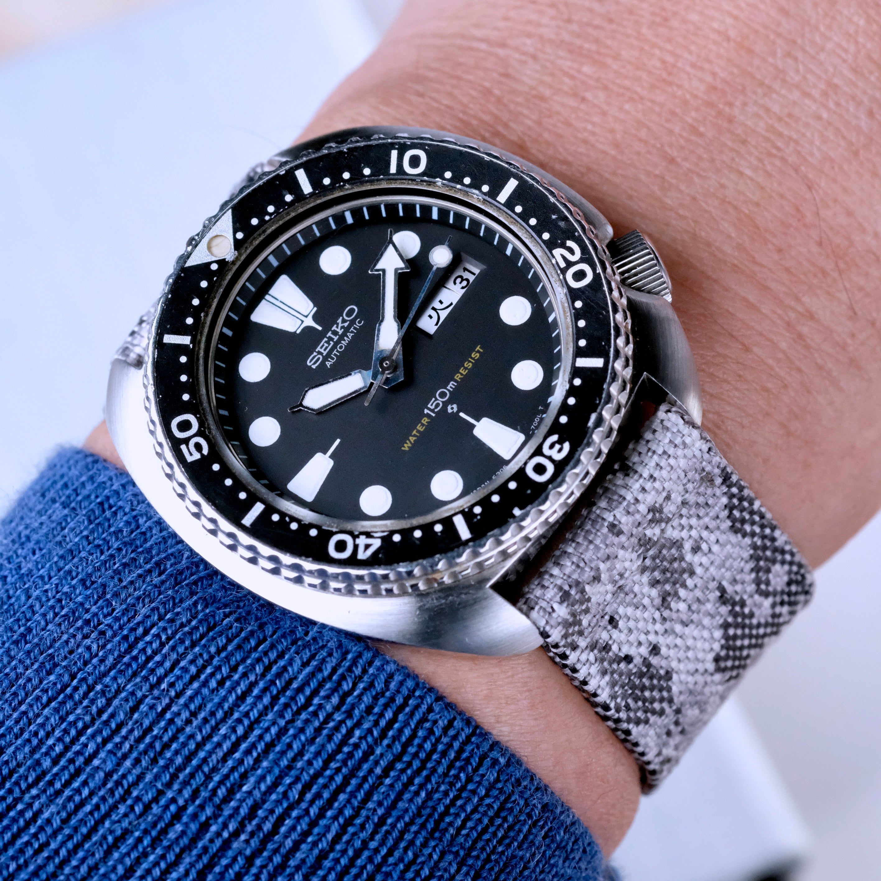 A Seiko dive watch with a black bezel, black dial, and white hour markers is worn on the wrist in a blue sleeve, paired with an RSM Watch Strap Snow Camo 2-Piece jacquard woven strap.