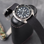 Close-up of VTG Mil in Black by RSM Watch Strap with a black fabric band, stainless steel buckle, and a dive watch on a leather case.