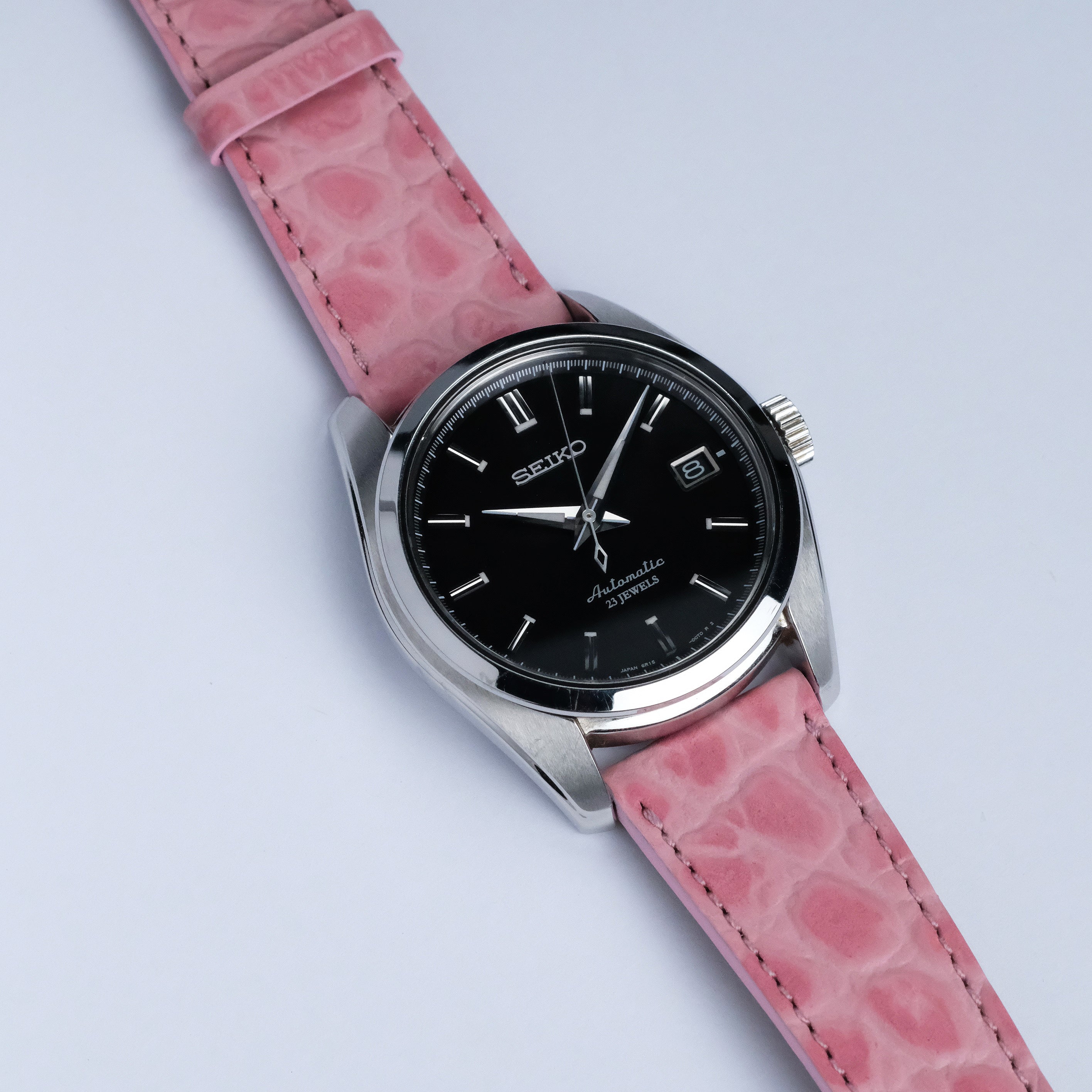 A Seiko wristwatch with a black dial, silver hour markers, and a date window is shown on a white background with an RSM Watch Strap Alligator Circular Grain Calf Leather Watch Strap in pink.