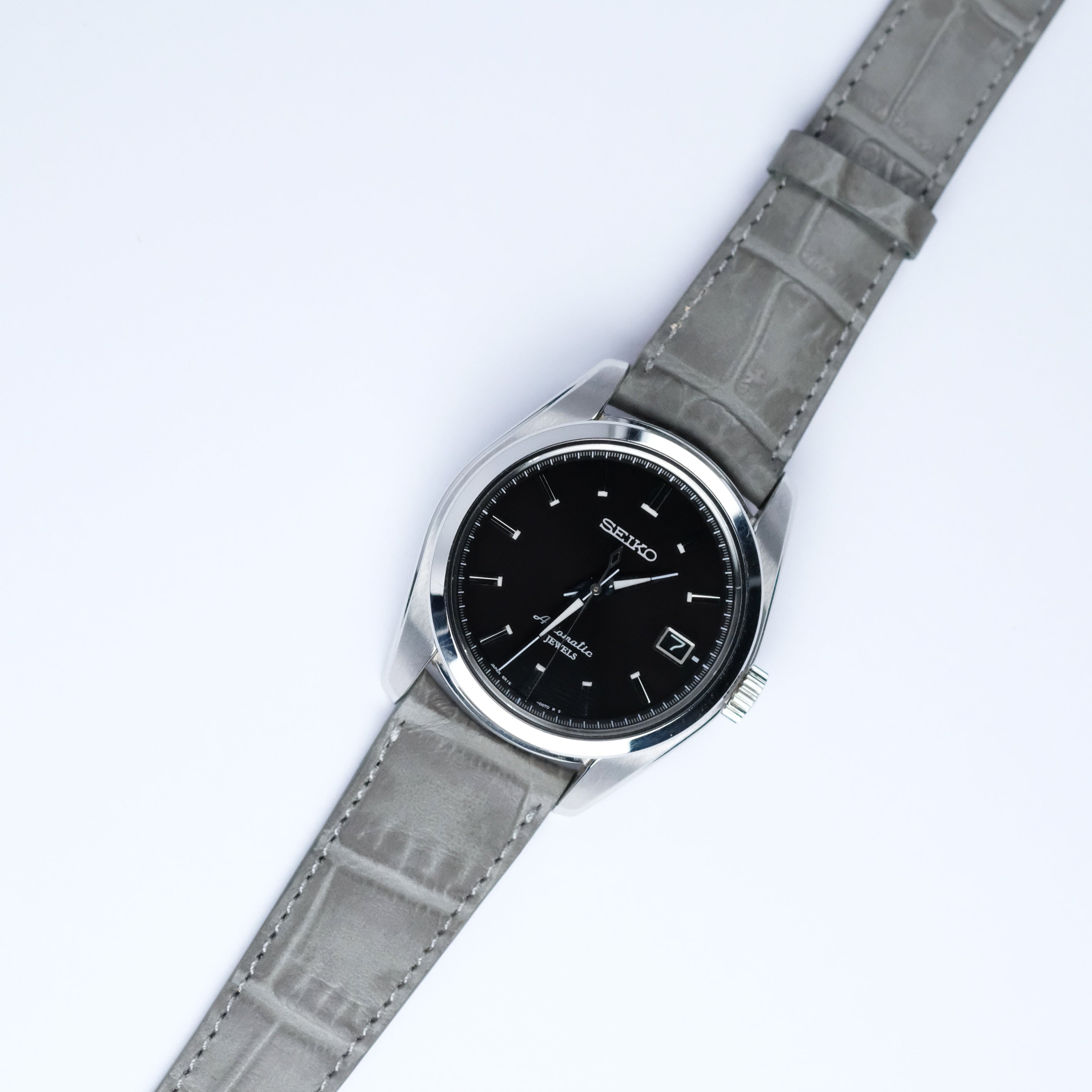 A Seiko watch with a black dial, silver markers, and date display is paired with the RSM Watch Strap Curved Lugs Alligator Rectangular Grain Watch Strap in Grey, highlighting Japanese craftsmanship on a white background.