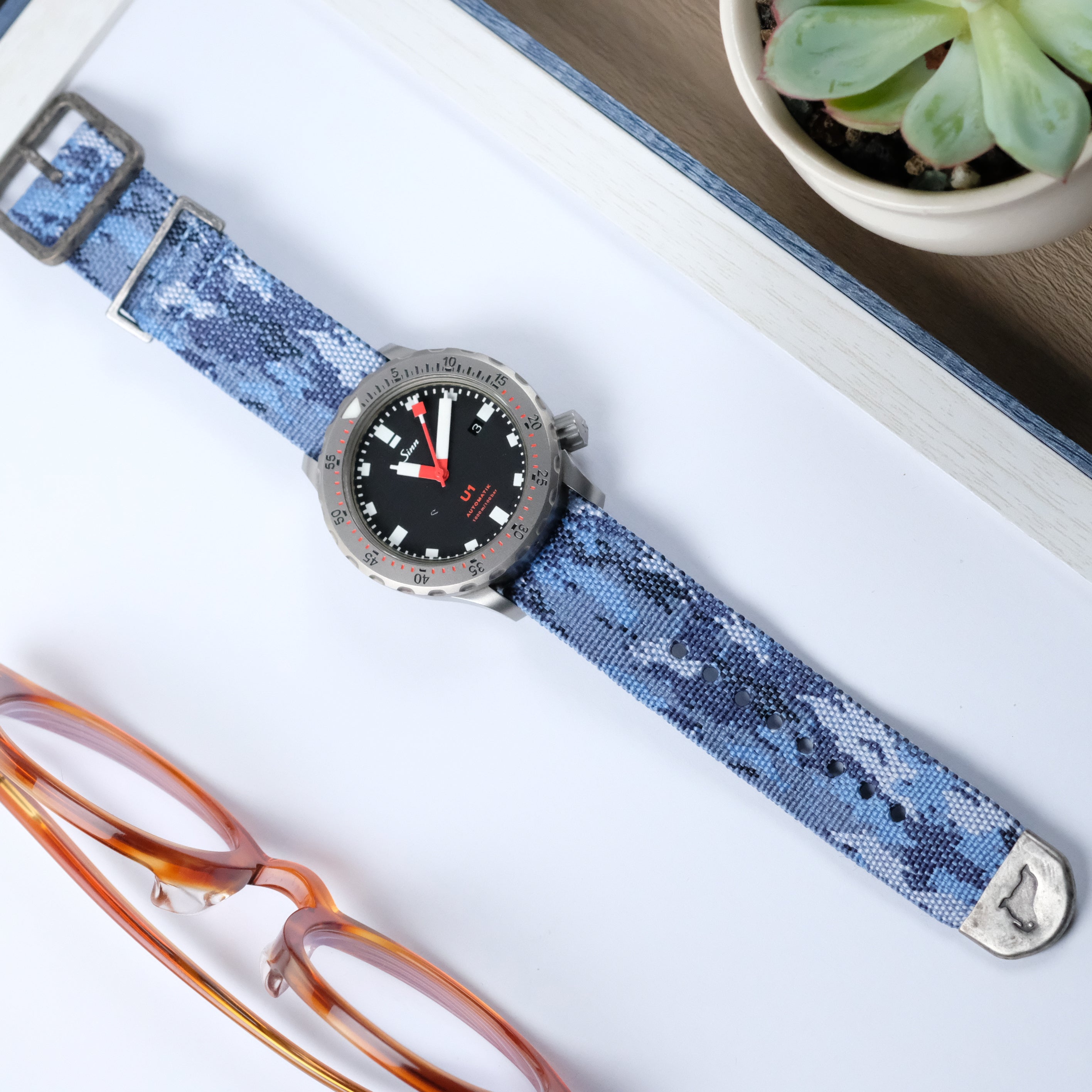 An RSM Watch Strap with an Ocean Camo Zulu Watch Strap lies on a white surface beside orange eyeglasses and a small potted succulent.