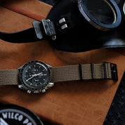 The VTG Mil in Green by RSM Watch Strap with a khaki fabric strap and stainless steel buckle rests on a brown surface next to aviator goggles.