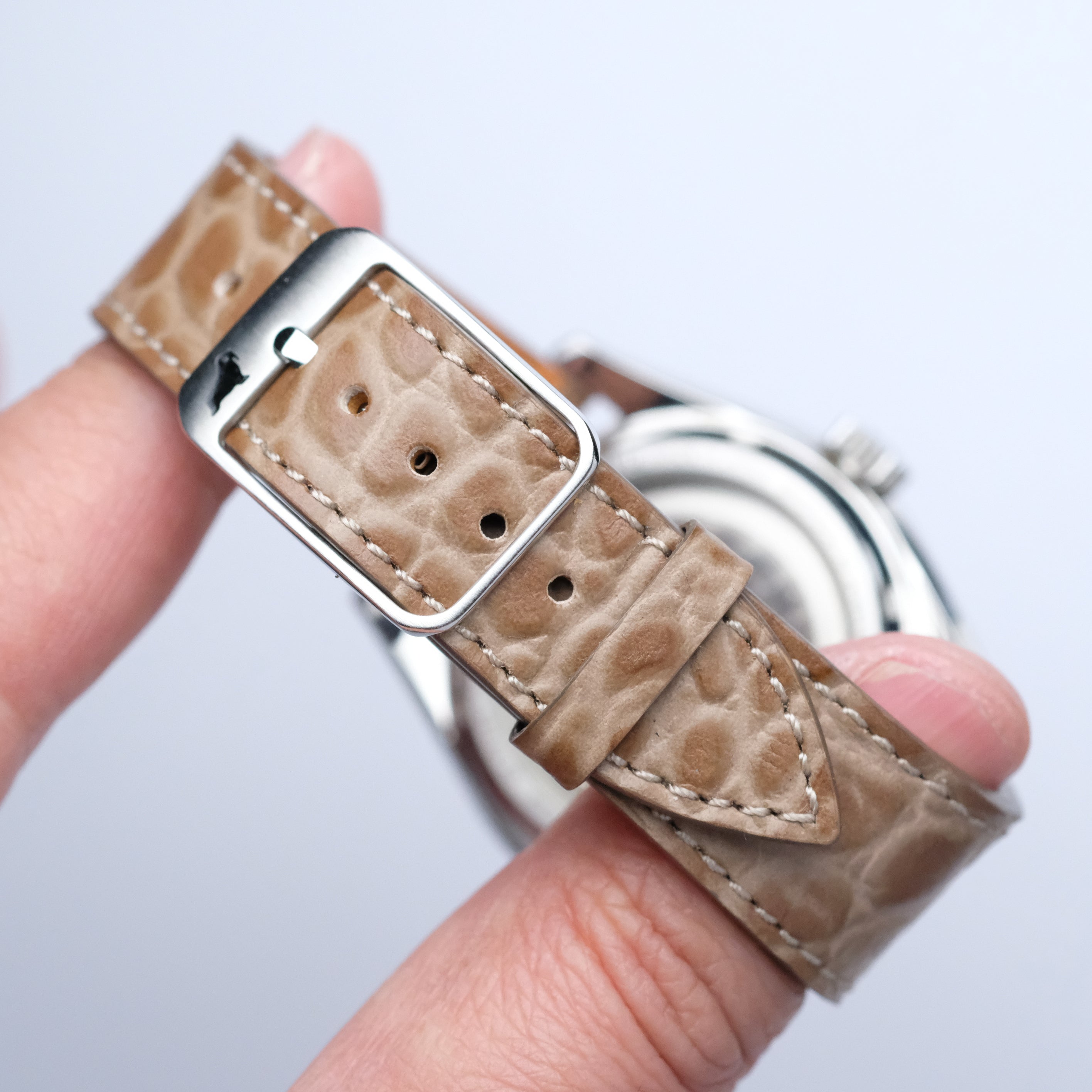 A hand displays the RSM Watch Strap’s Alligator Circular Grain Calf Leather Watch Strap in Taupe, its detailed stitching and fine texture inspired by Japanese craftsmanship, with a blurred watch face highlighting the elegant band.