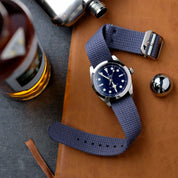 The VTG Mil in Blue by RSM Watch Strap, with its blue fabric strap and dark blue face, rests on leather beside a flask and metal object.