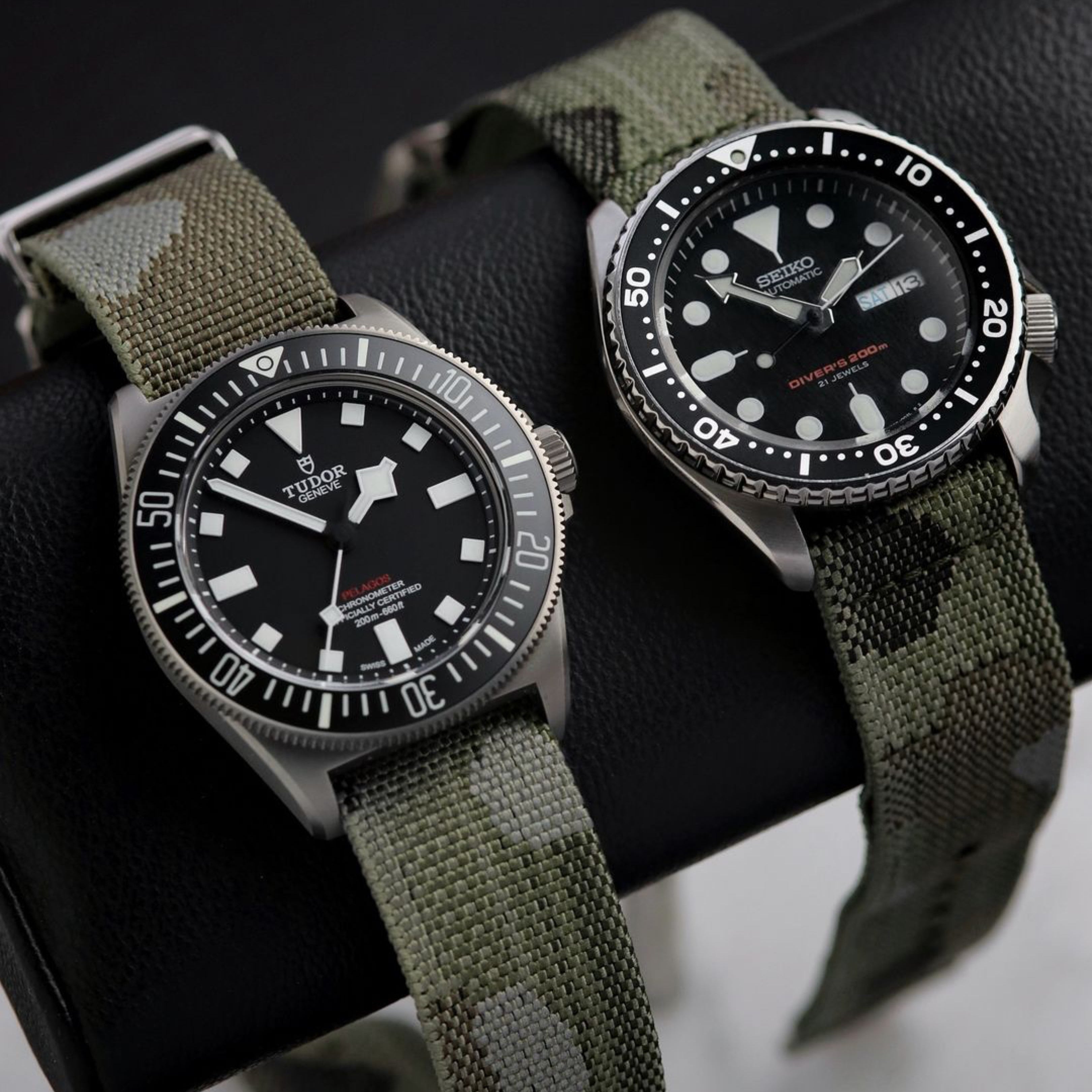 Two wristwatches with RSM Watch Strap's Camo 2-Piece straps are displayed on a black leather holder. The left watch is a Tudor, and the right is a Seiko with a black dial and bold white markers.