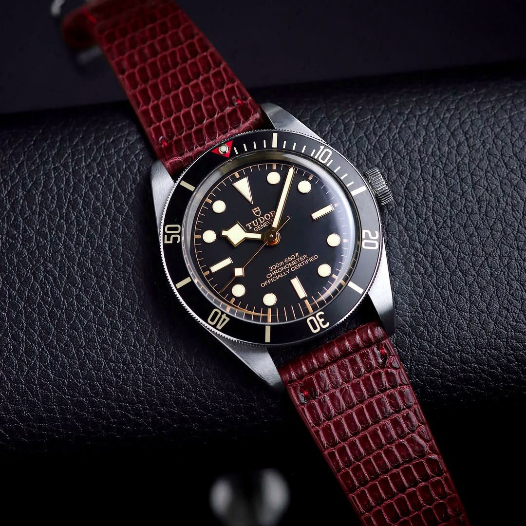 A Tudor wristwatch with a black dial and bezel is displayed on a textured black surface, paired with the RSM Watch Strap Lizard Leather Watch Strap Vertical Grain in Red for an elegant touch.