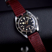 A Tudor wristwatch with a black dial and bezel is displayed on a textured black surface, paired with the RSM Watch Strap Lizard Leather Watch Strap Vertical Grain in Red for an elegant touch.