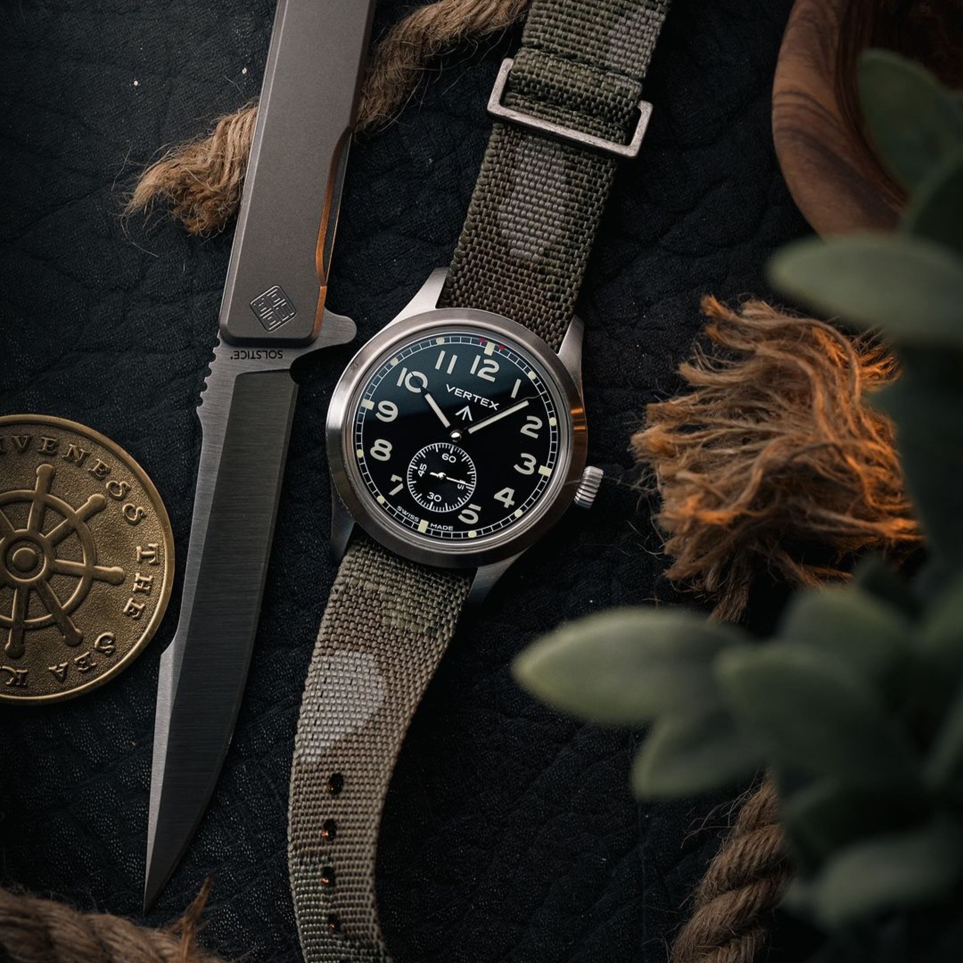 The RSM Watch Strap Camo Zulu Watch Strap is displayed on a wristwatch beside a tactical knife, nautical coin, and rope on a dark surface, with green leaves partly visible in the foreground.