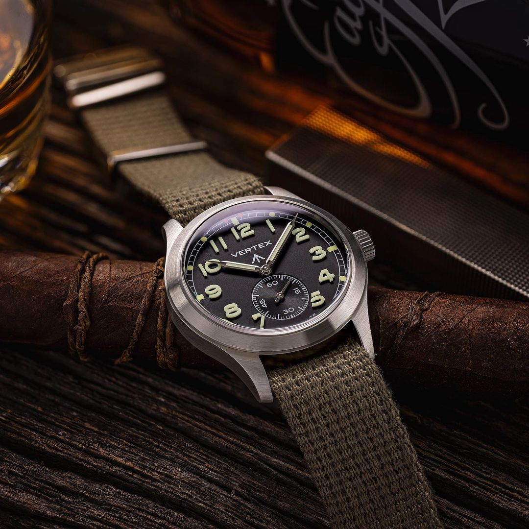 Vertex M100A resting on a chair with RSM Vintage Military polycotton single pass strap, adjustable length. Background: whisky bottle, glasses, vintage lighter.