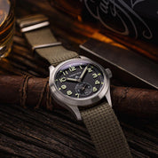 Vertex M100A resting on a chair with RSM Vintage Military polycotton single pass strap, adjustable length. Background: whisky bottle, glasses, vintage lighter.