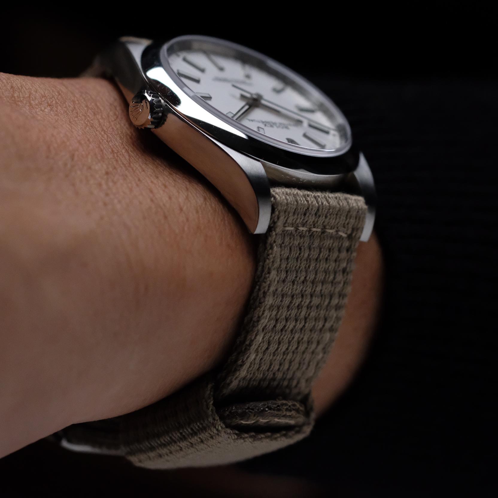 A close-up of a wrist wearing a silver watch with a white face and the RSM Watch Strap Vintage Military 2-Piece Watch Strap in Steel, featuring quick-release spring bars and a textured beige fabric, against a dark background.