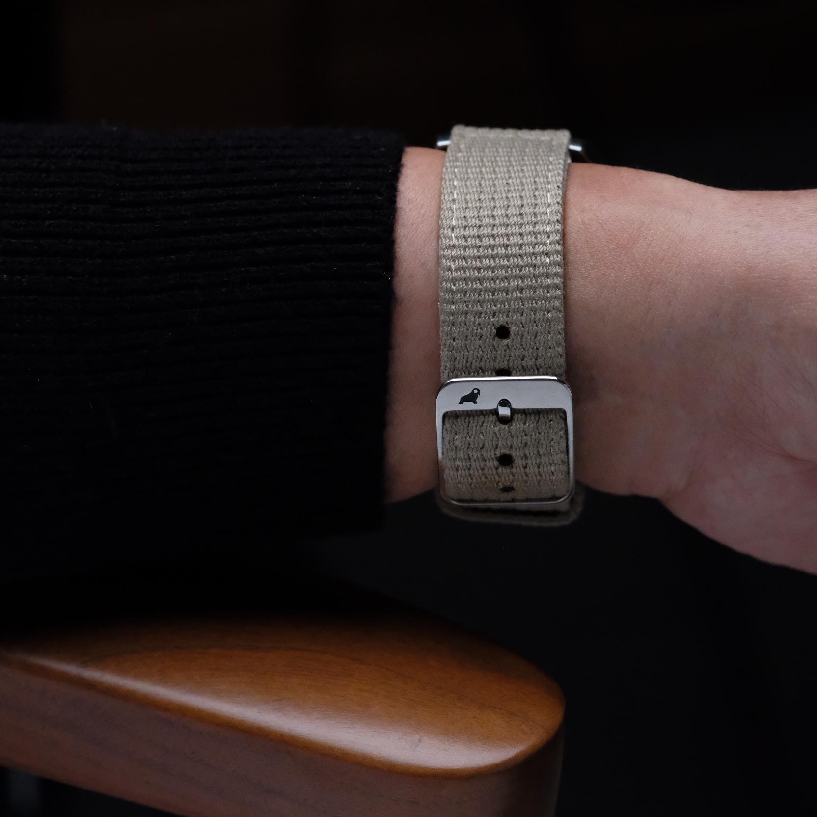 A close-up of a wrist wearing the RSM Watch Strap Vintage Military 2-Piece Watch Strap in Steel, Apple Watch-compatible with a silver buckle, rests on a wooden armrest. The person wears a black ribbed long-sleeve top.