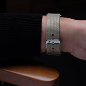 A close-up of a wrist wearing the RSM Watch Strap Vintage Military 2-Piece Watch Strap in Steel, Apple Watch-compatible with a silver buckle, rests on a wooden armrest. The person wears a black ribbed long-sleeve top.