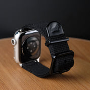 A close-up of the Vintage Military 2-Piece Watch Strap in Black by RSM Watch Strap, an Apple Watch-compatible polycotton band with a metal clasp embossed with a dog logo, shown on a wooden surface against a black background.