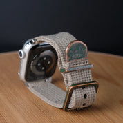 The RSM Watch Strap Vintage Military 2-Piece in Green, compatible with Apple Watch, features a beige woven fabric band and weathered bronze buckle, displayed on wood against a black background.