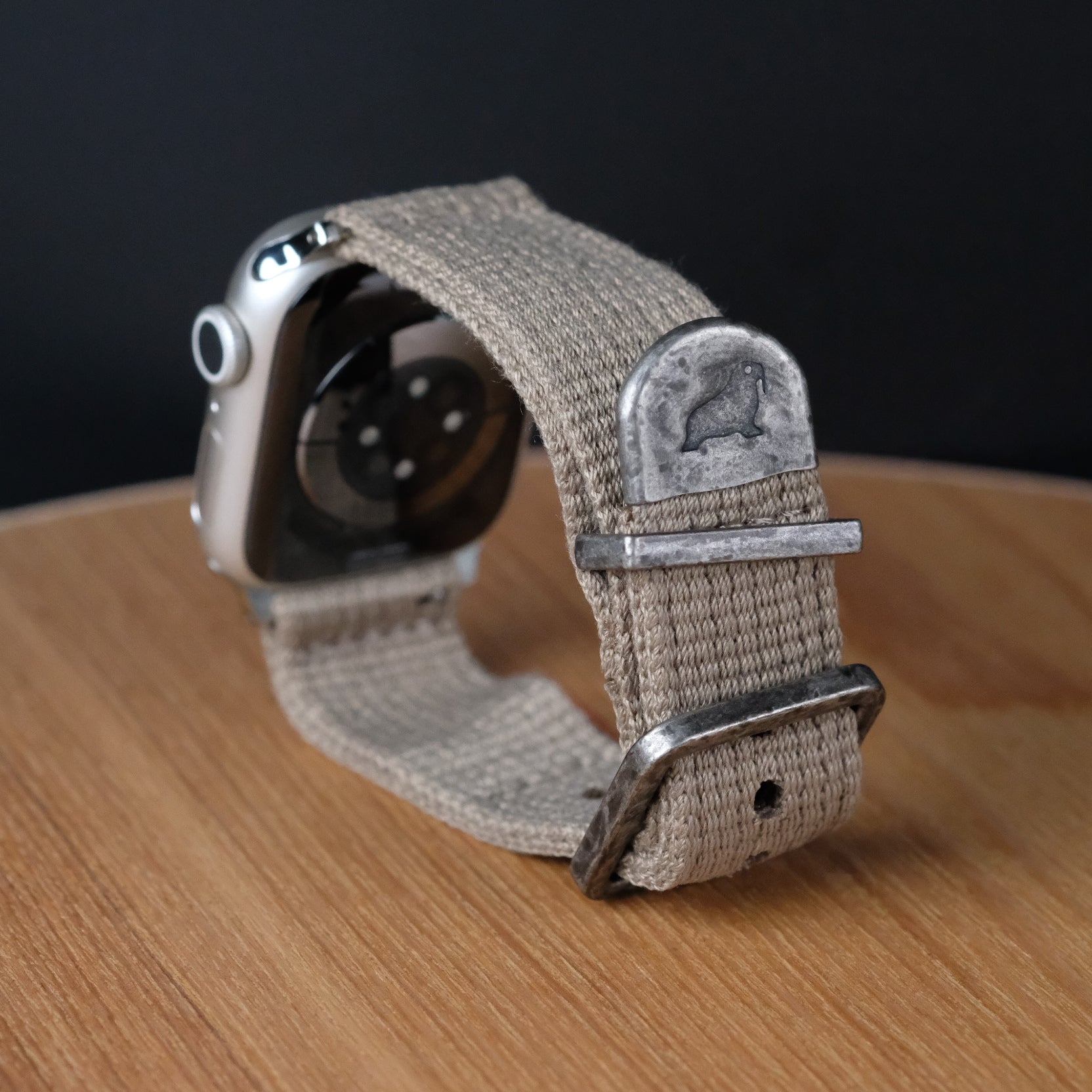 The RSM Watch Strap Vintage Military 2-Piece Watch Strap in Khaki, compatible with Apple Watch, features a metal clasp and stamped animal design, shown on a wooden surface with a dark background.