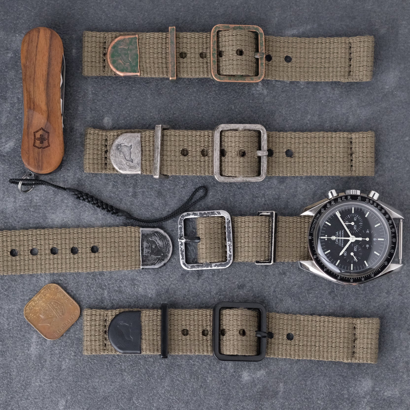 Poly cotton woven green vintage style watch band in Distressed bronze with green patina, distressed silver, distressed black and matte black hardware. Swiss army wooden small tool knife, vintage square coin. Omega Speedmaster. RSM Walrus logo on end caps of distressed hardware.