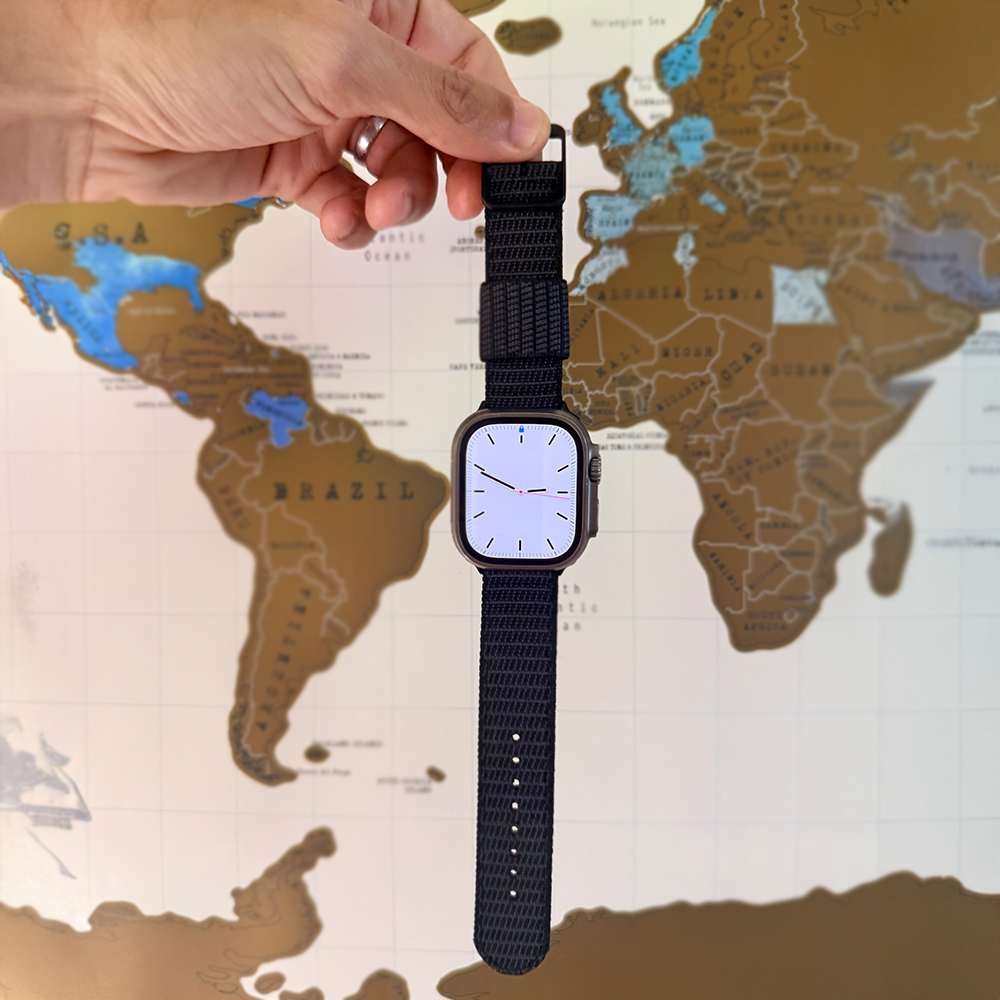 A hand holds an X-Hatch 2-Piece Watch Strap in Black by RSM Watch Strap, attached to an Apple Watch-compatible smartwatch with a blank screen, positioned in front of a world map with brown, blue, and white countries.