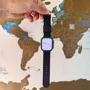 A hand holds an X-Hatch 2-Piece Watch Strap in Black by RSM Watch Strap, attached to an Apple Watch-compatible smartwatch with a blank screen, positioned in front of a world map with brown, blue, and white countries.