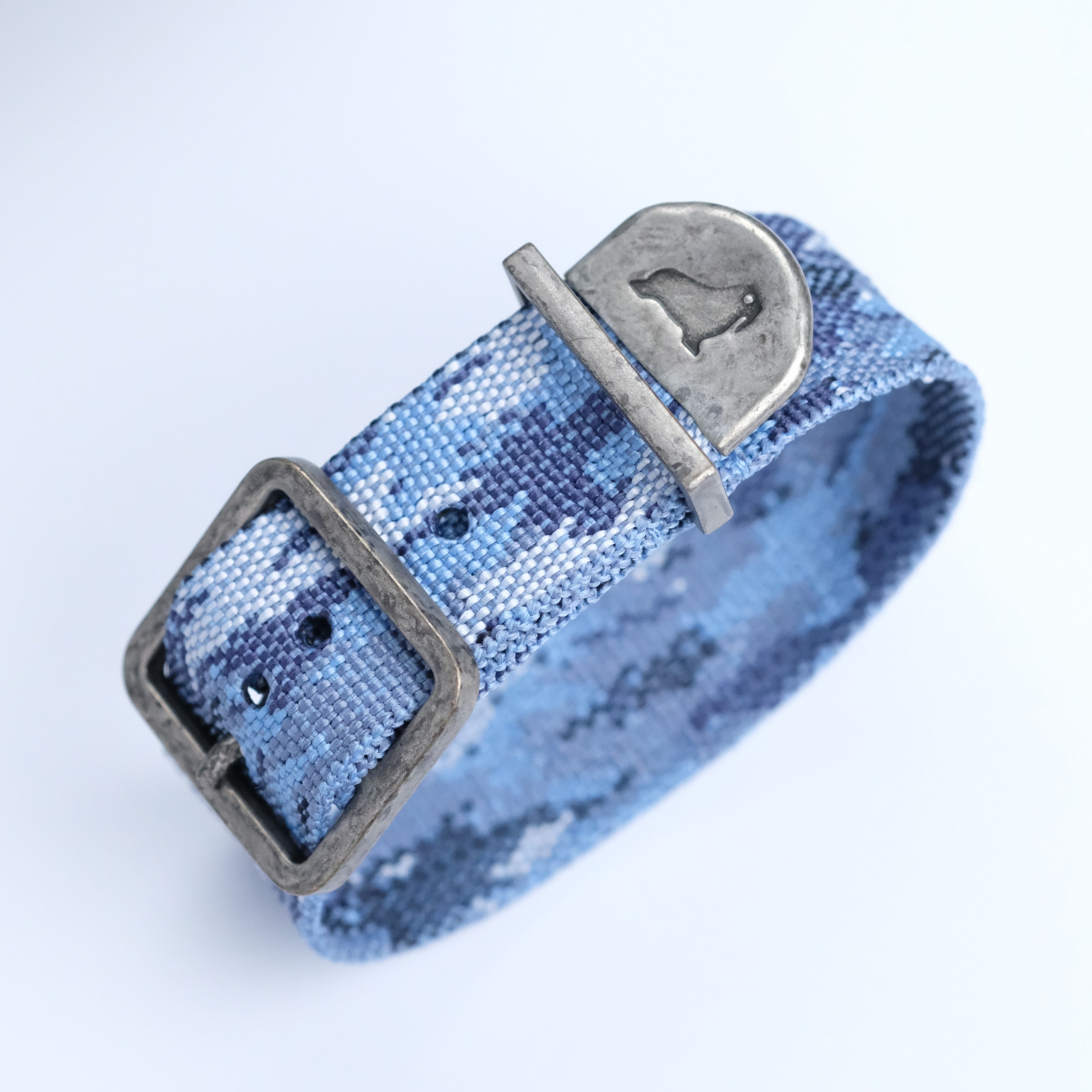 The RSM Watch Strap Ocean Camo Zulu Watch Strap is a jacquard-woven blue bracelet with a worn silver rectangular buckle and an engraved penguin silhouette on a metallic plate, displayed on a white background.