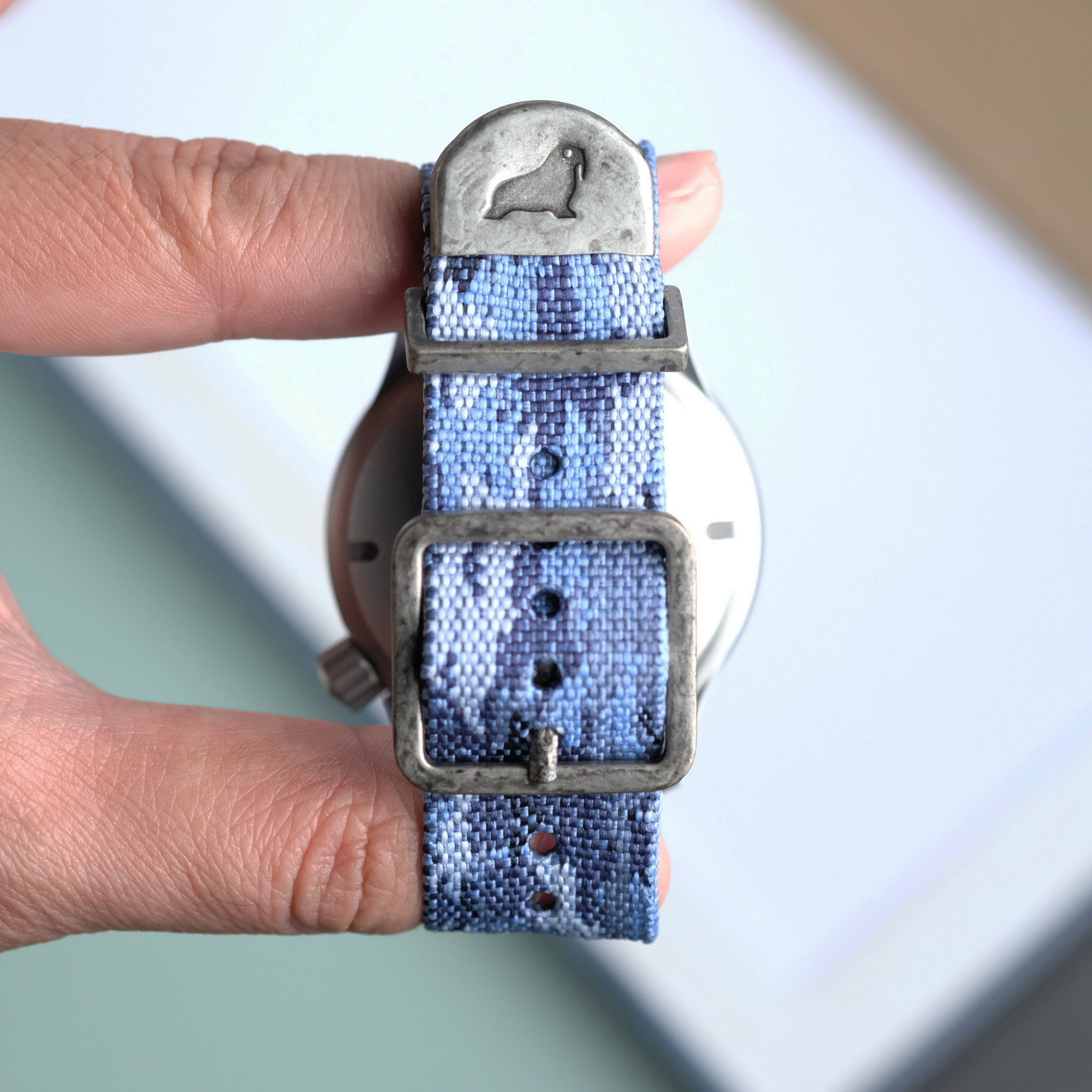 A hand holds an RSM Watch Strap Ocean Camo Zulu Watch Strap with a metal buckle and an engraved sea lion on the strap’s end. The background is softly blurred in light colors.