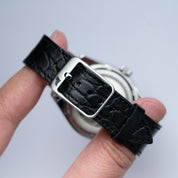 A hand holds the RSM Watch Strap’s Alligator Circular Grain Calf Leather Watch Strap in Black, featuring a silver buckle and detailed Japanese craftsmanship, set against a plain white background.