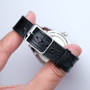 A hand holds the RSM Watch Strap Alligator Grain Calf Leather Watch Strap in Black with a silver buckle, showcasing its craftsmanship against a plain white background.