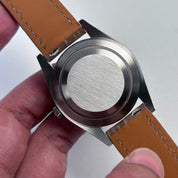 Close-up of a watch with a brown leather strap held by a hand on a light gray background