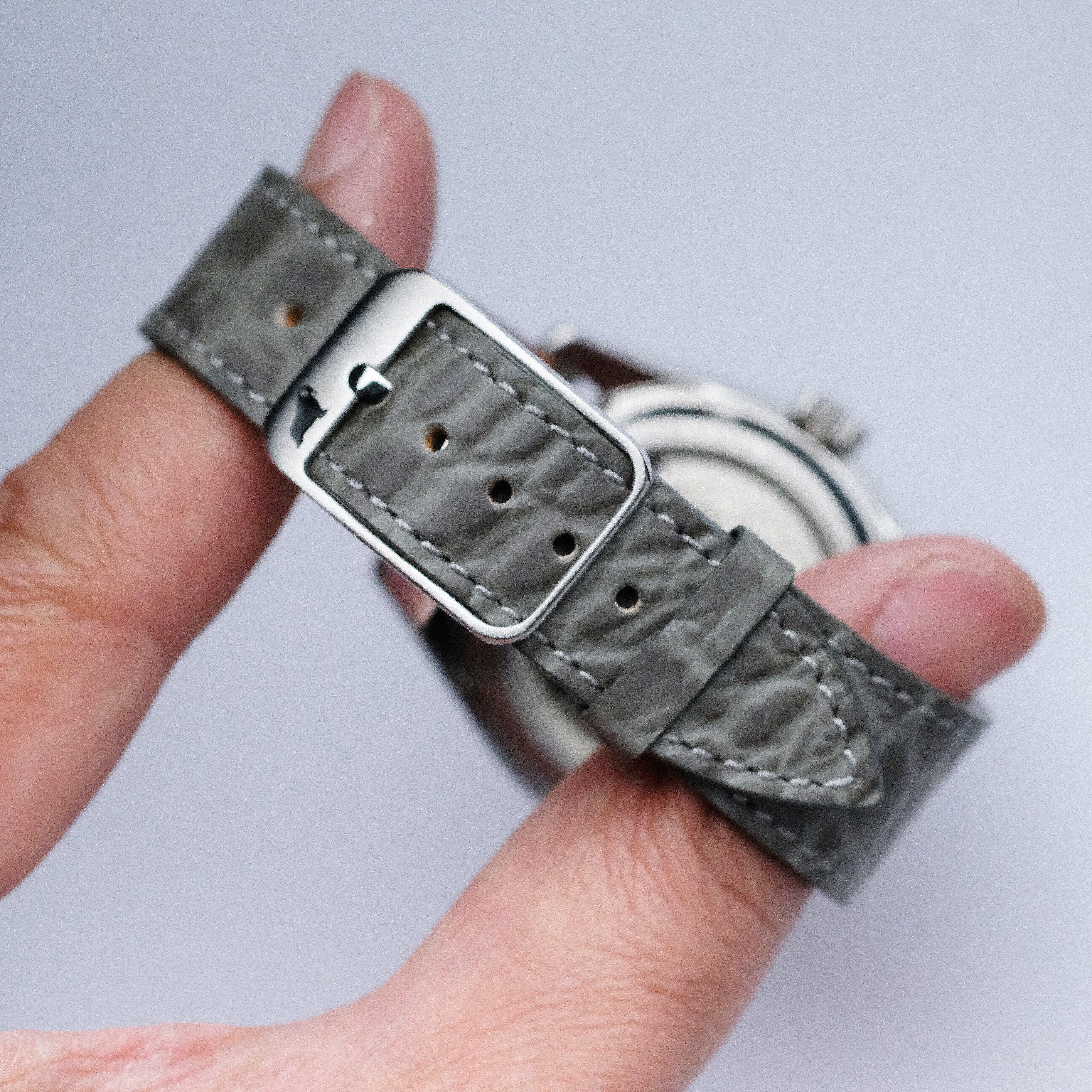 A hand holds the RSM Watch Strap Alligator Circular Grain Calf Leather Watch Strap in Grey, featuring a silver buckle and showcasing its detailed Japanese craftsmanship on the back.