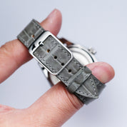 A hand holding the RSM Watch Strap Alligator Grain Calf Leather Watch Strap in Grey with a silver buckle, shown from the back against a plain light background, highlights enduring Japanese craftsmanship.