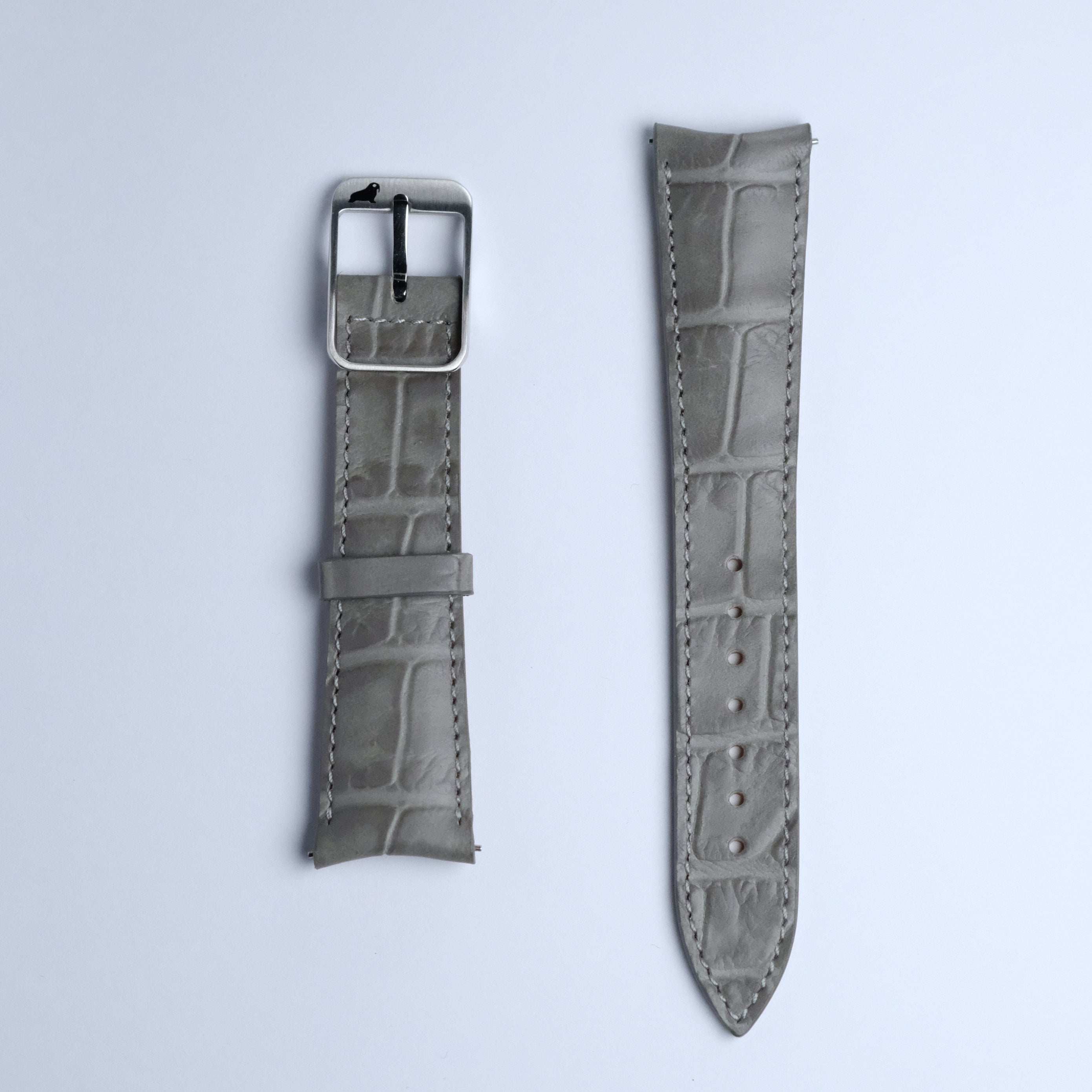 The Curved Lugs Alligator Rectangular Grain Watch Strap in Grey by RSM Watch Strap, shown in two flat pieces—one with a metal buckle and one with adjustment holes—is crafted from embossed calf leather on a white background.