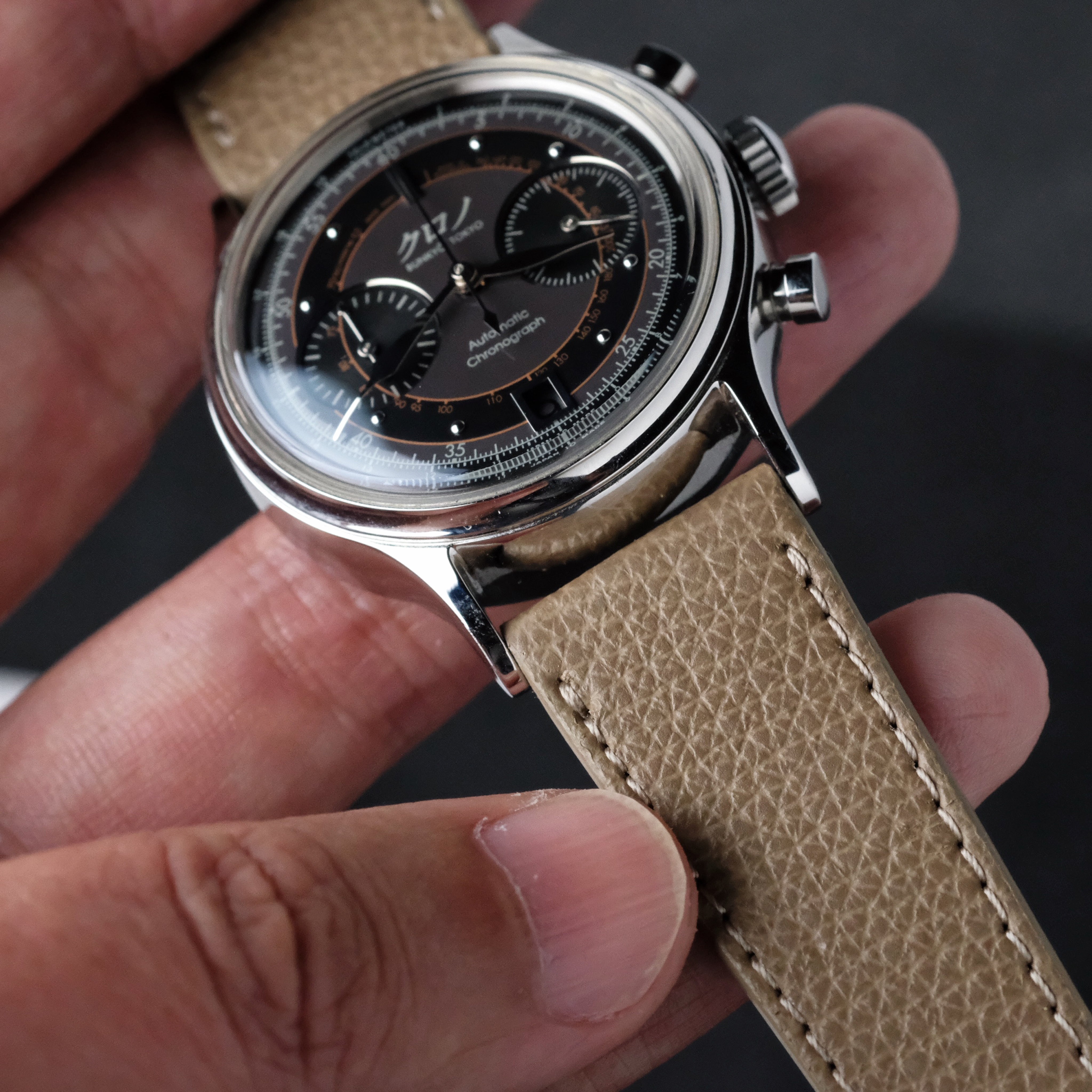 A close-up of a hand holding a wristwatch with a black dial, three subdials, silver casing, and chronograph pushers paired with the Italian Epsom Leather Watch Strap in Beige by RSM Watch Strap.