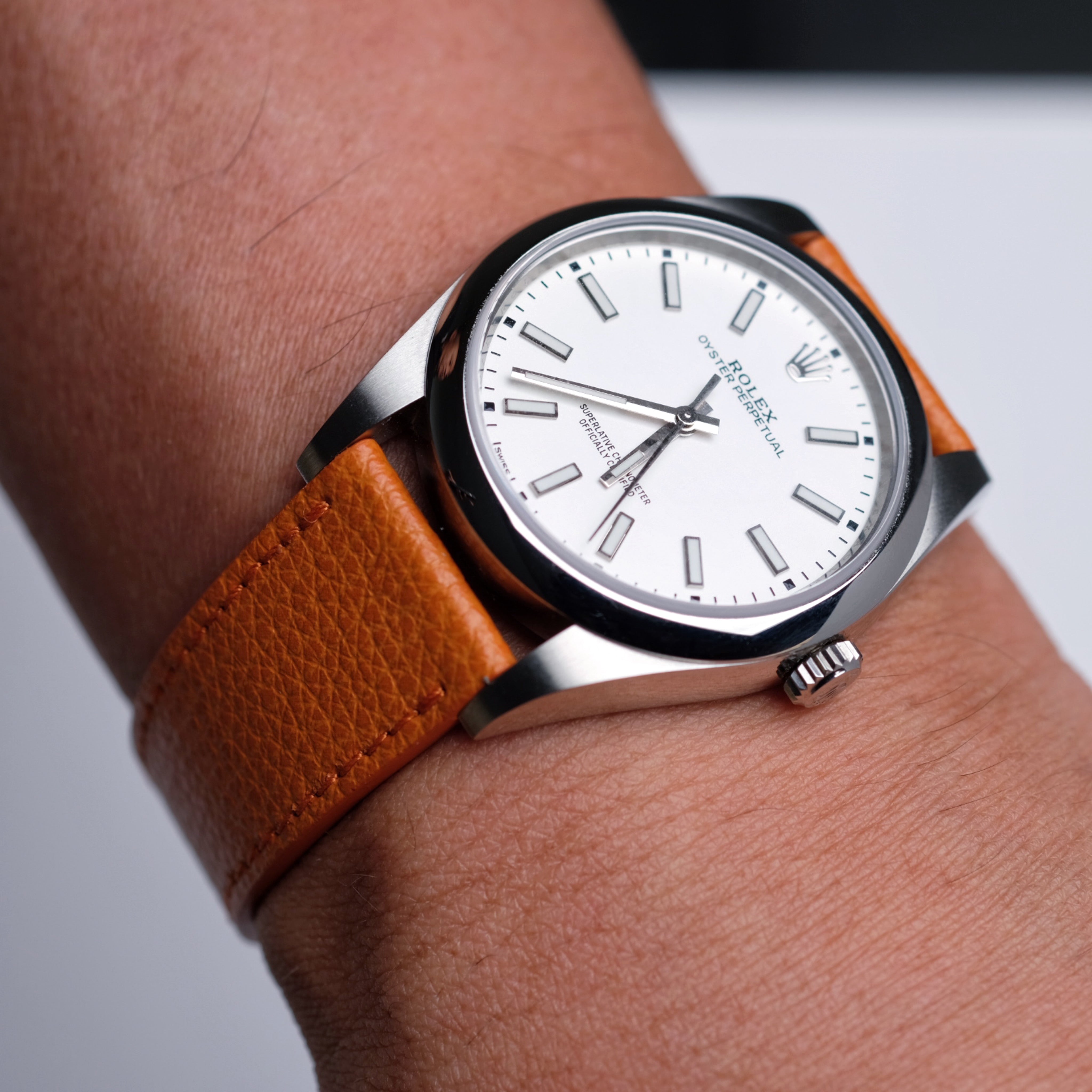A close-up of a wrist wearing a Rolex Oyster Perpetual with a white face and silver markers, paired with the RSM Watch Strap Italian Epsom Leather Watch Strap in Orange.