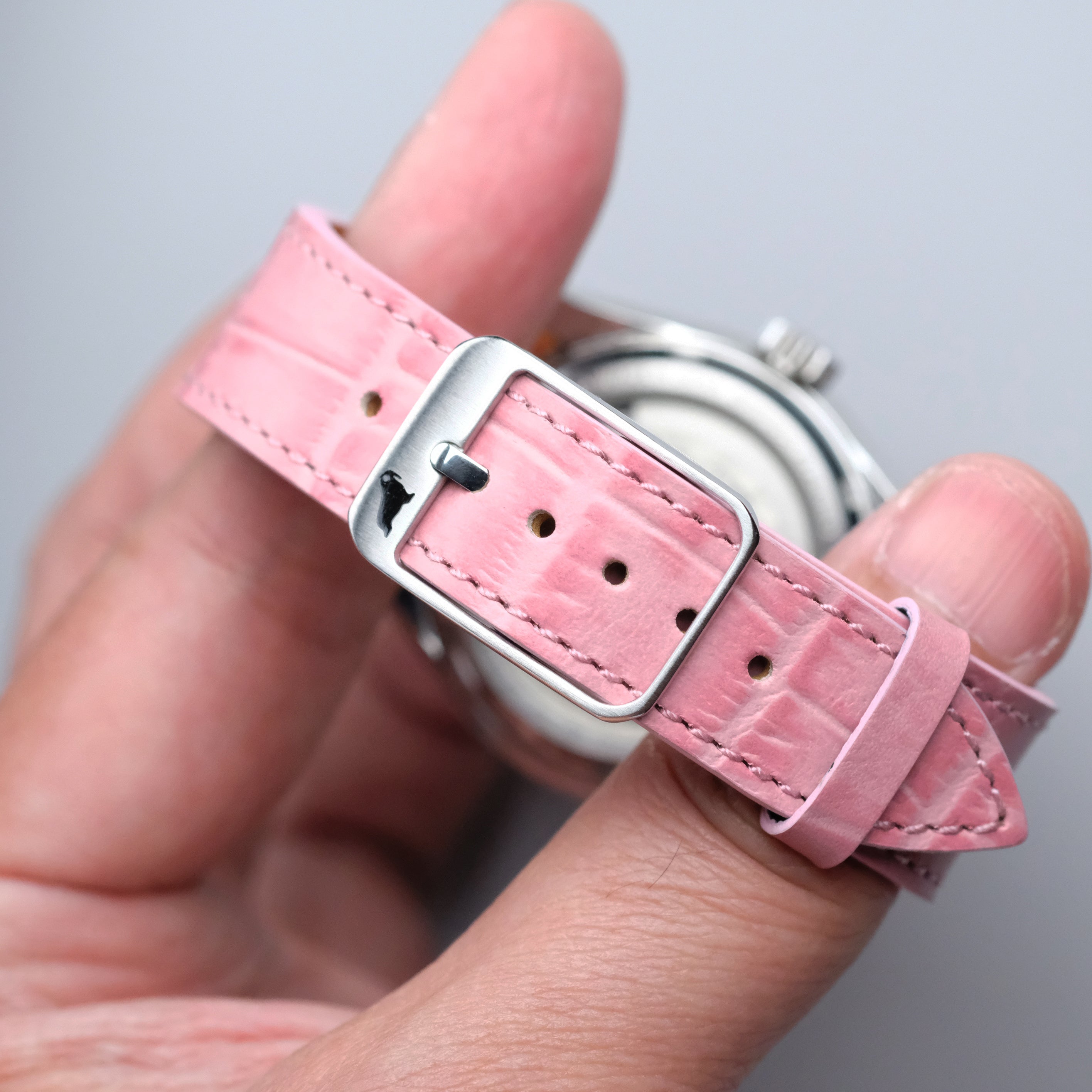 A hand displays the RSM Watch Strap Curved Lugs Alligator Circular Grain Watch Strap in Pink, featuring an embossed alligator leather texture and a silver buckle. The strap and buckle highlight Japanese craftsmanship with a blurred watch face.