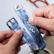 A person holds the Ocean Camo 2-Piece Watch Strap by RSM Watch Strap, displaying its jacquard-woven polyester material, holes, and metal clasp. Another hand holds the buckle end against a blurred background with a plant.