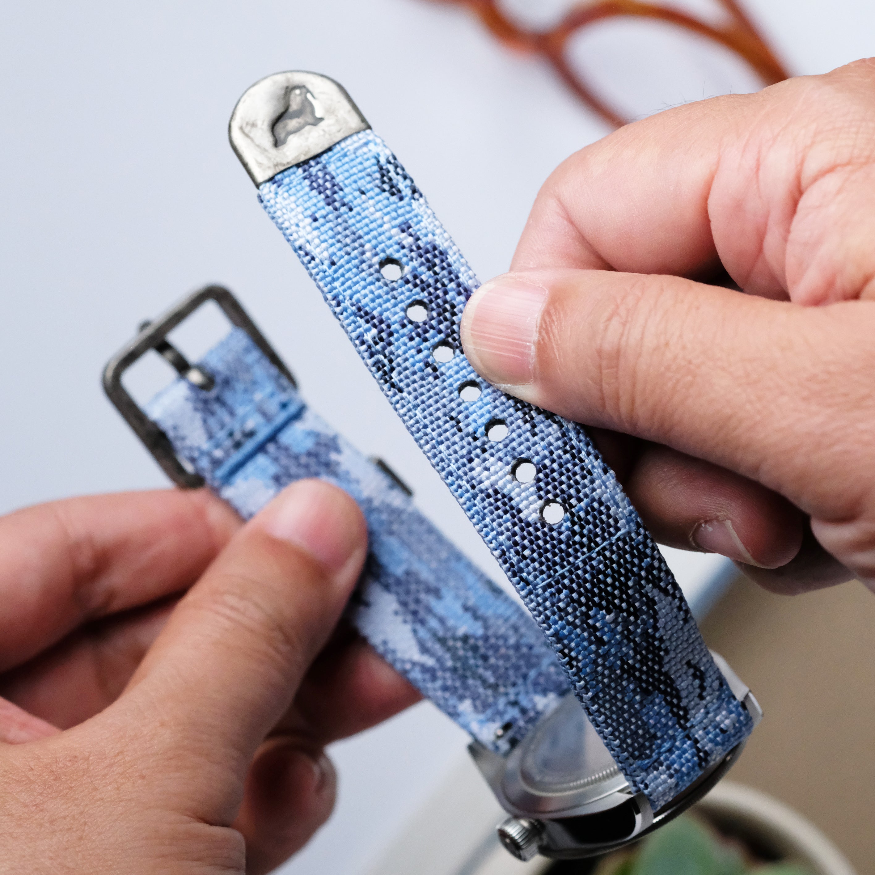 A person holds the Ocean Camo 2-Piece Watch Strap by RSM Watch Strap, displaying its jacquard-woven polyester material, holes, and metal clasp. Another hand holds the buckle end against a blurred background with a plant.
