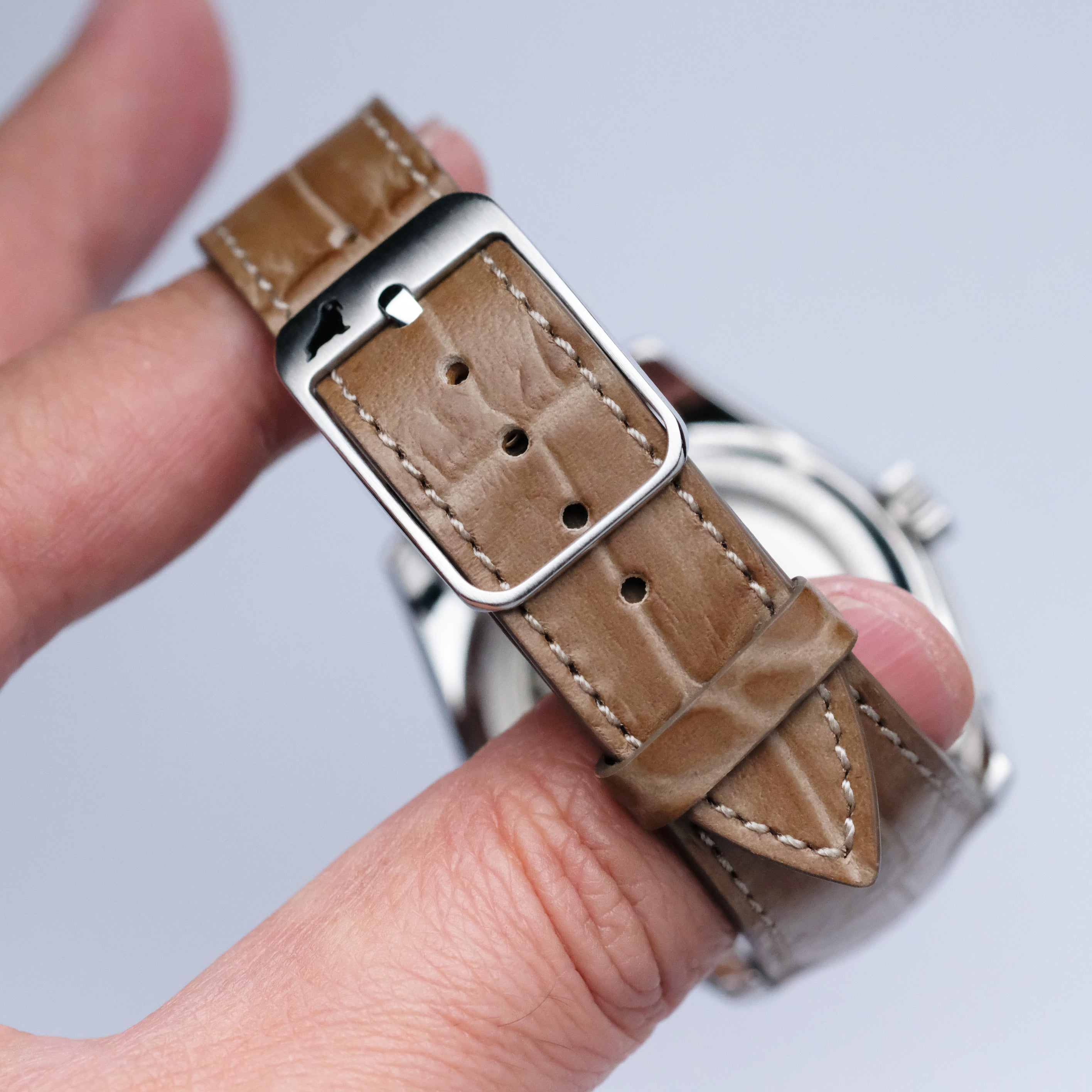 A close-up of a hand holding an RSM Watch Strap Alligator Grain Calf Leather Watch Strap in Taupe with visible stitching and a silver buckle, showcasing Japanese craftsmanship. The watch face is turned away from the camera.