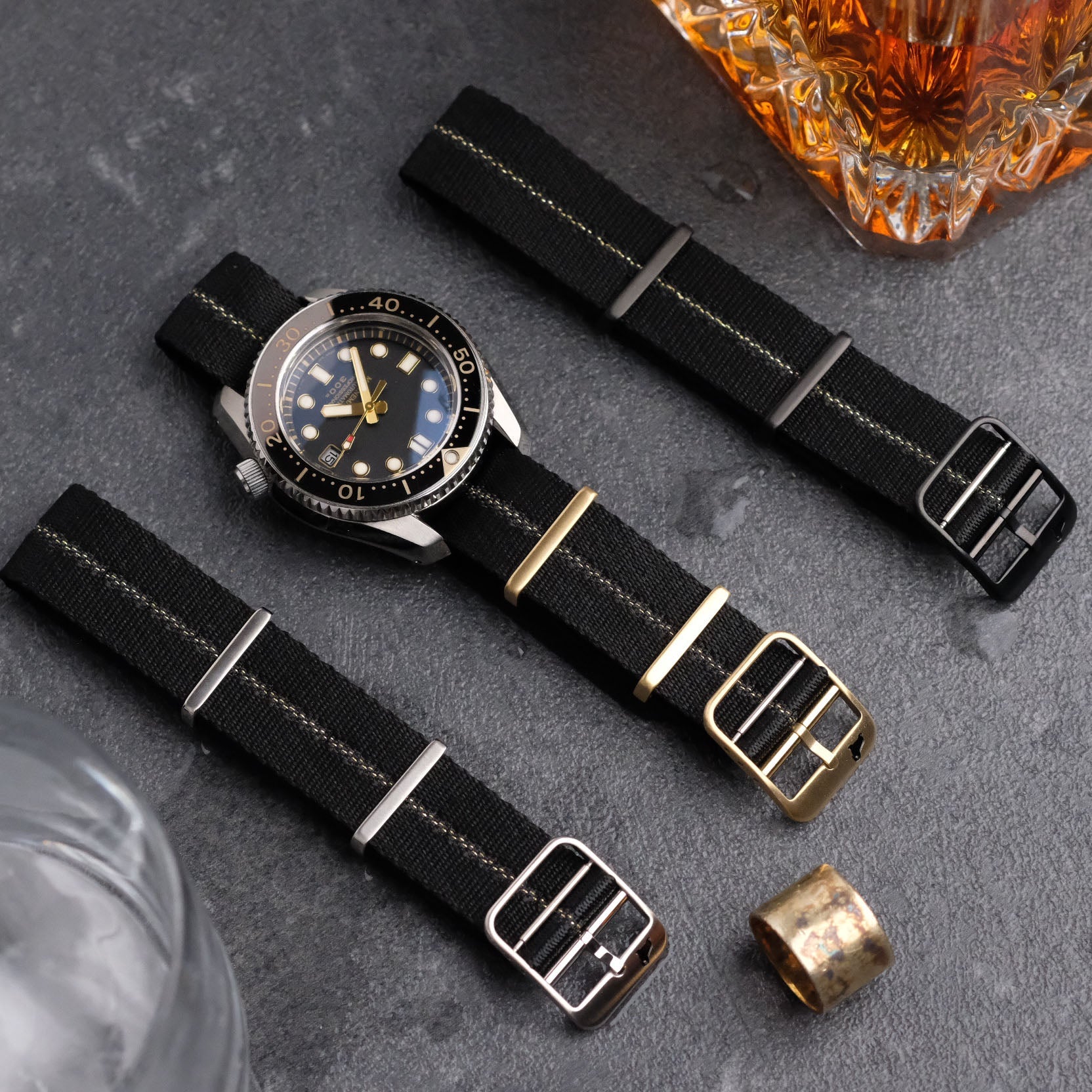 Luxurious nylon strap displayed on a dark surface with three black straps of varying buckle finishes, accented by a glass container and metal object.