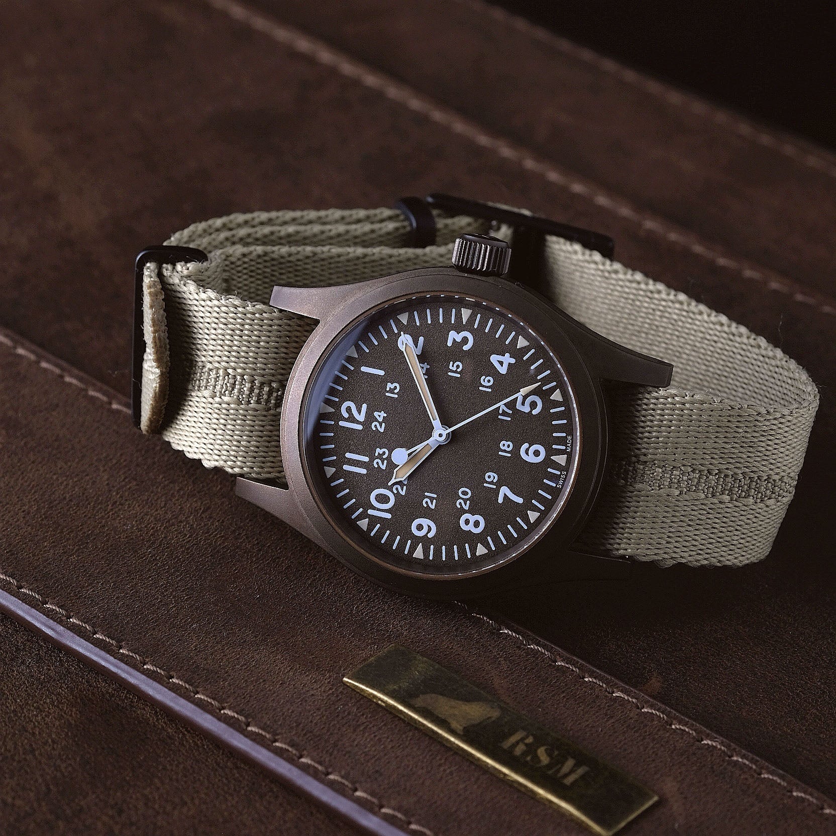 The Engraved Stripe in Stealth Khaki by RSM, featuring a brown bezel and jacquard weave khaki strap, rests on a dark brown textured surface.