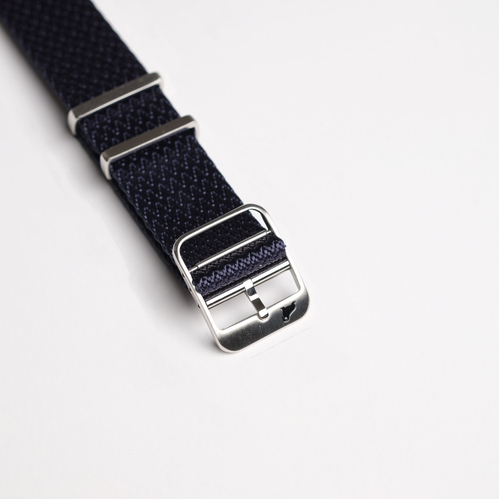 A close-up of the Herringbone Twill in Icarus Blue NATO watch band by RSM, featuring silver hardware, a metal buckle, and loops for securing excess length, laid against a white background.