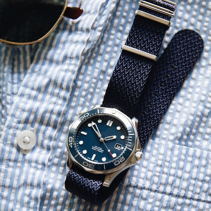 A luxury watch with a blue face and silver bezel rests on an RSM Watch Strap Herringbone Twill in Icarus Blue, with sunglasses in the top left corner, exuding elegance.